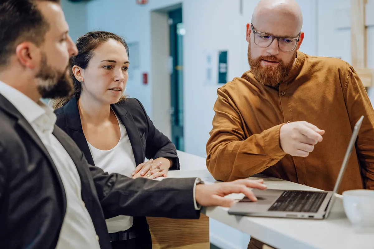 Three people in a business meeting discussing something on a laptop screen. One person is pointing at the screen while the others listen attentively.
