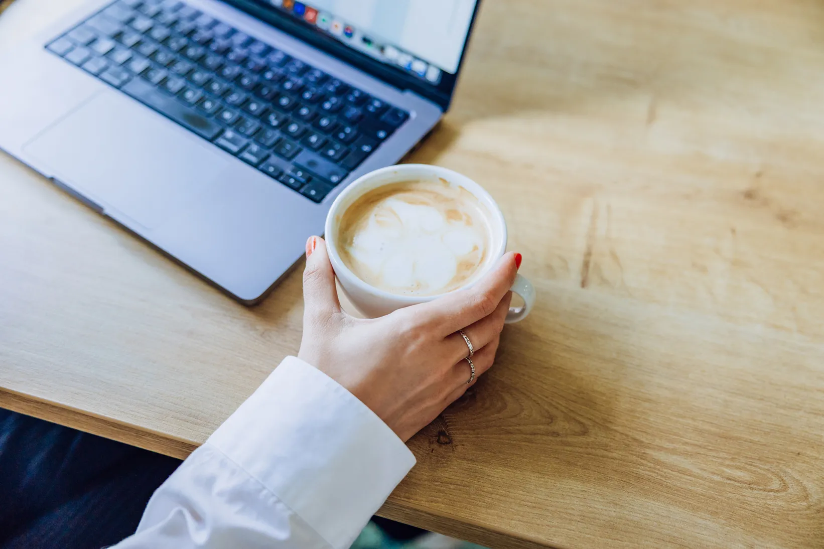 A person holding a cup of coffee next to an open laptop on a wooden table.