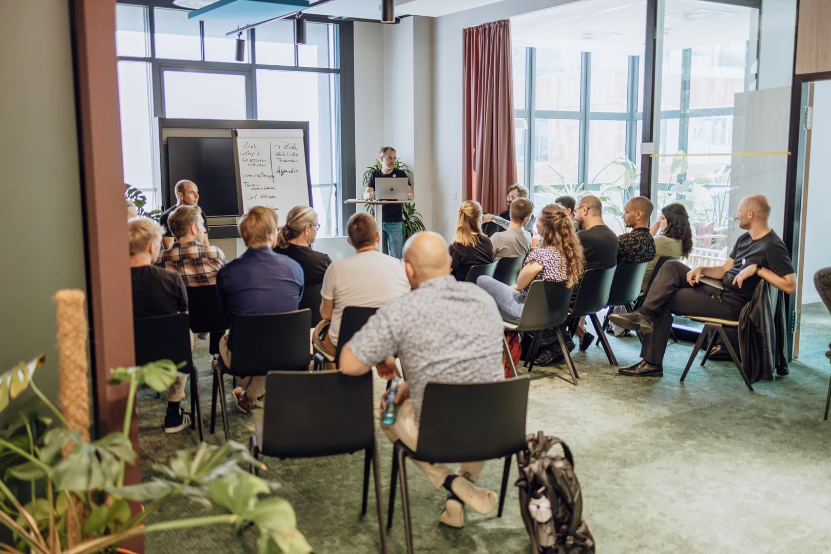 A group of people seated in a conference room, attentively listening to a speaker standing at the front with a laptop and a whiteboard displaying notes.