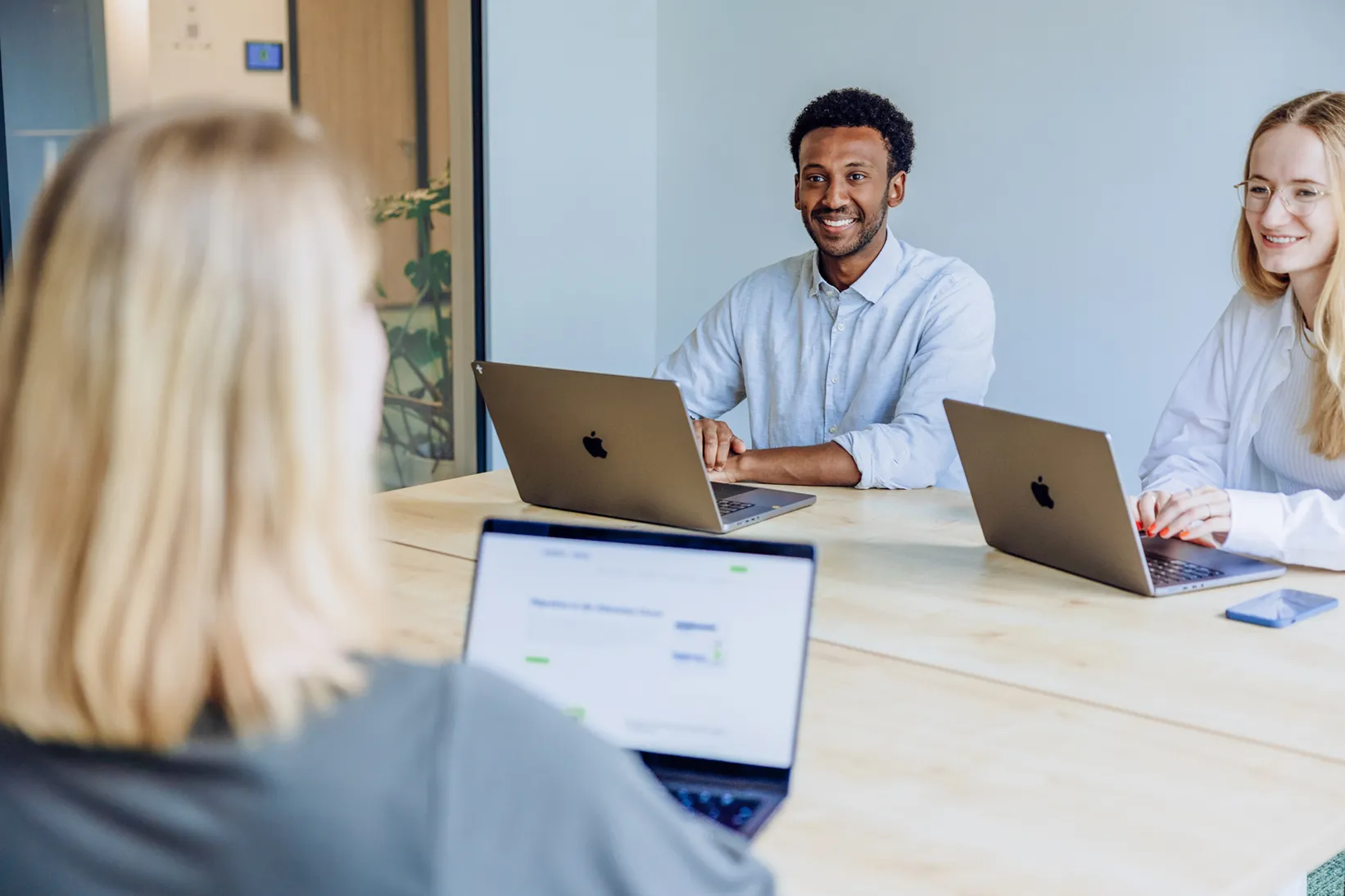 Three people sitting at a table with laptops, engaged in a friendly office meeting. They are smiling and appear to be discussing something on their screens.