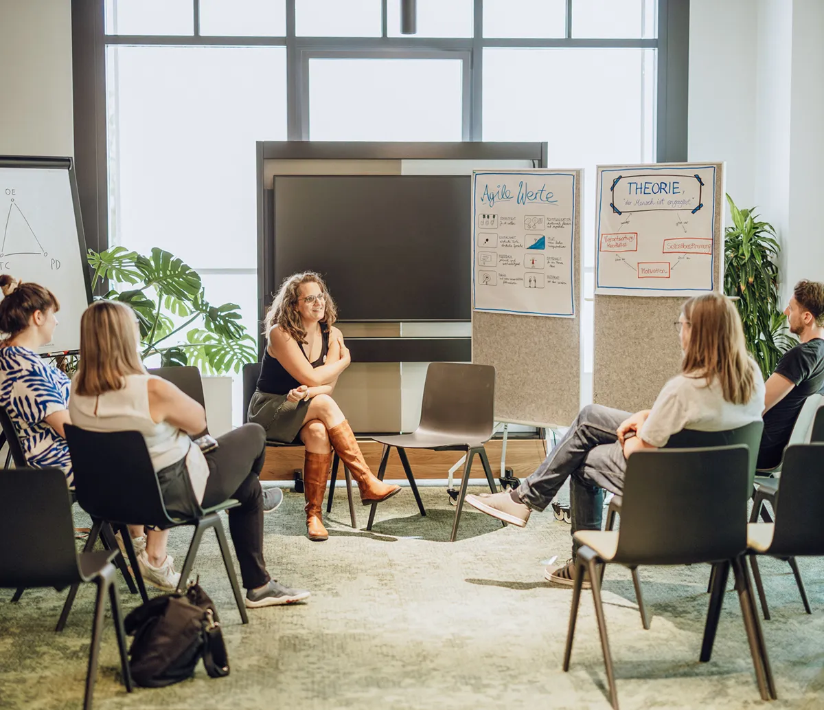 A group of people sitting in a circle in a bright room, engaged in a discussion. There are flipcharts with diagrams and notes in the background.