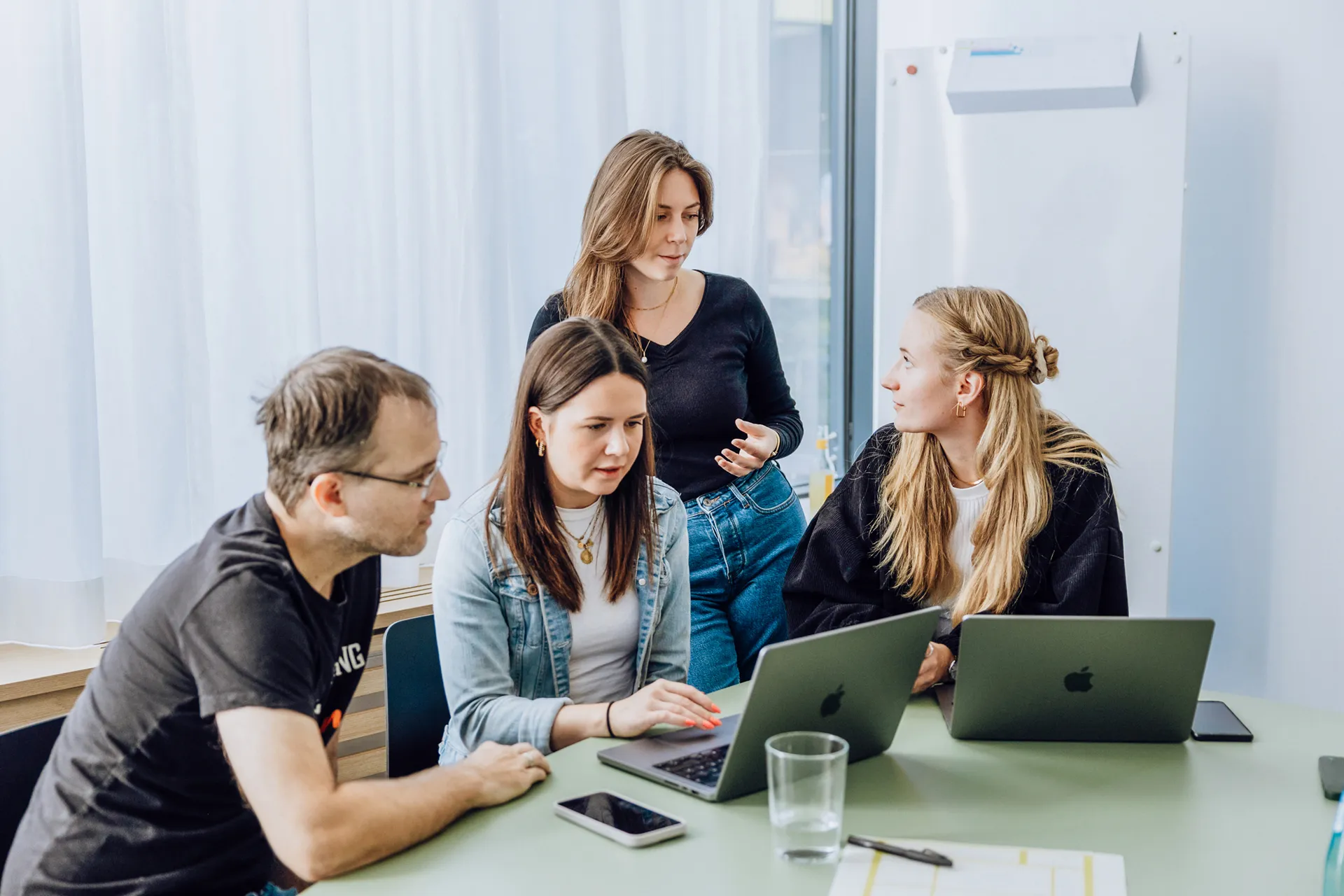 Four people gathered around a table with laptops, engaged in a discussion. One person is standing, while the others are seated, focusing on the laptops.