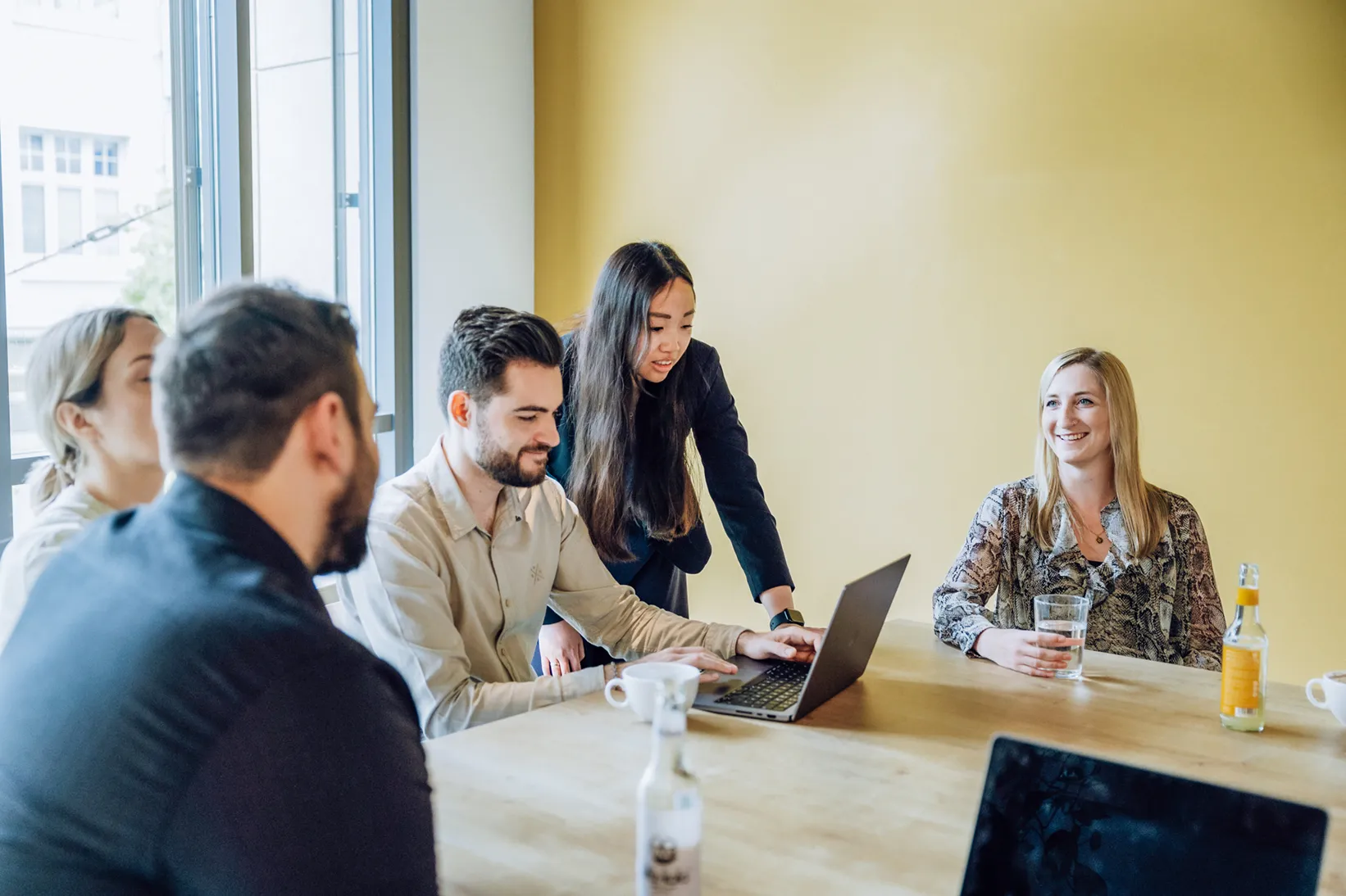 A group of people sitting around a table with laptops, engaged in a discussion. One person is standing and pointing at a laptop screen, while others are seated, smiling and holding drinks. The setting is a bright room with a yellow wall.