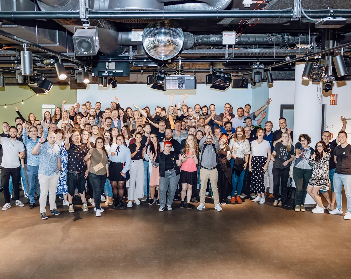 A large group of people posing together indoors, smiling and waving at the camera. The setting appears to be a modern office or event space with a disco ball on the ceiling.