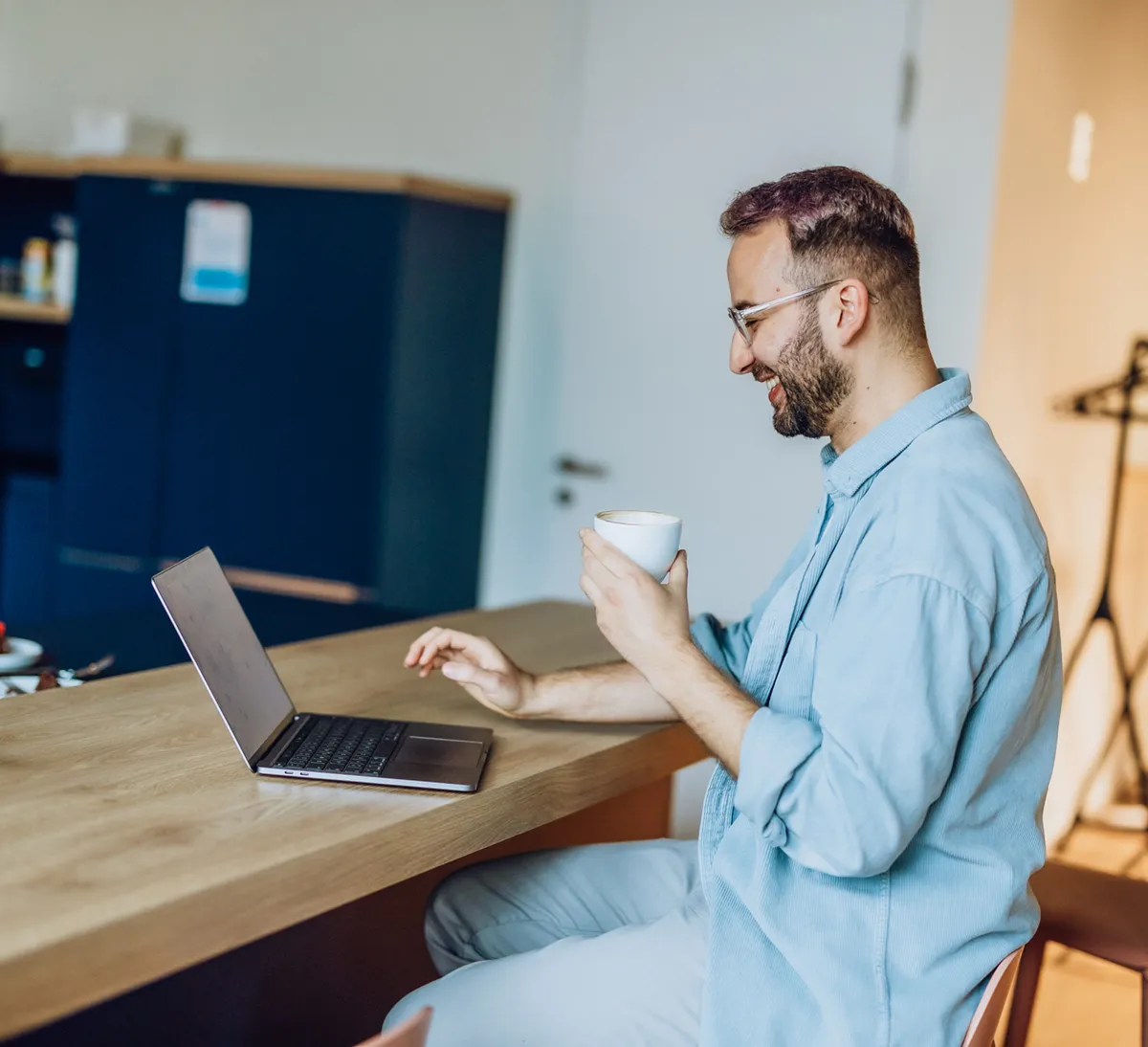 Man sitting at a kitchen counter with a laptop and holding a coffee cup, smiling while looking at the screen.