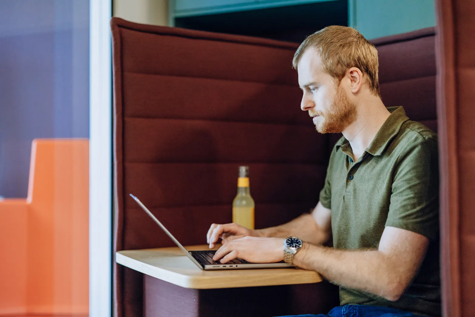 Man working on a laptop in a cozy booth with a drink on the table.