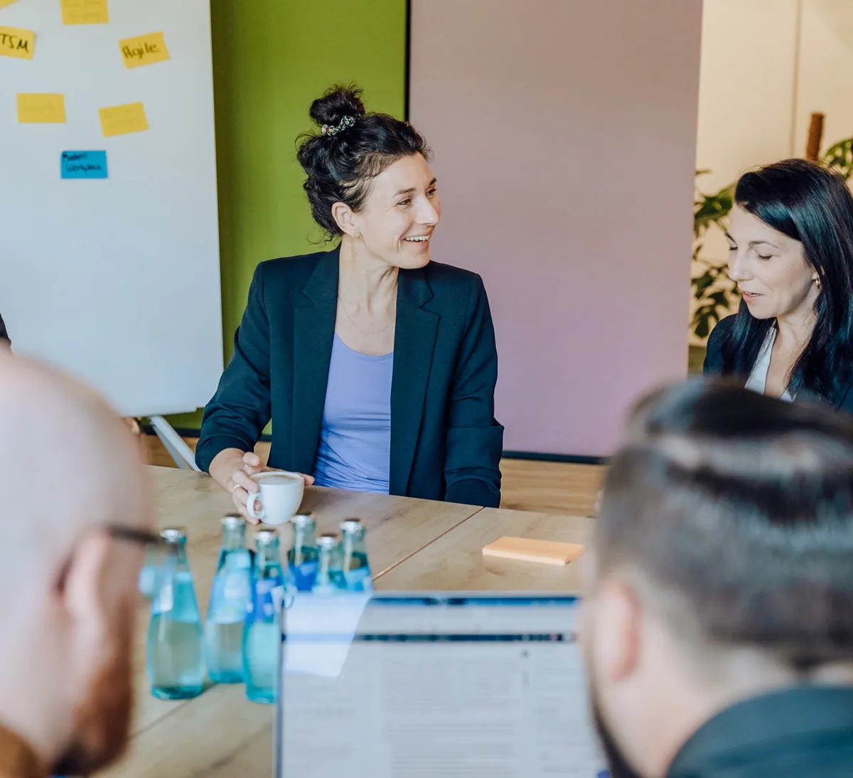 Group of people in a meeting room with a flipchart and sticky notes, discussing around a table with water bottles and a laptop.
