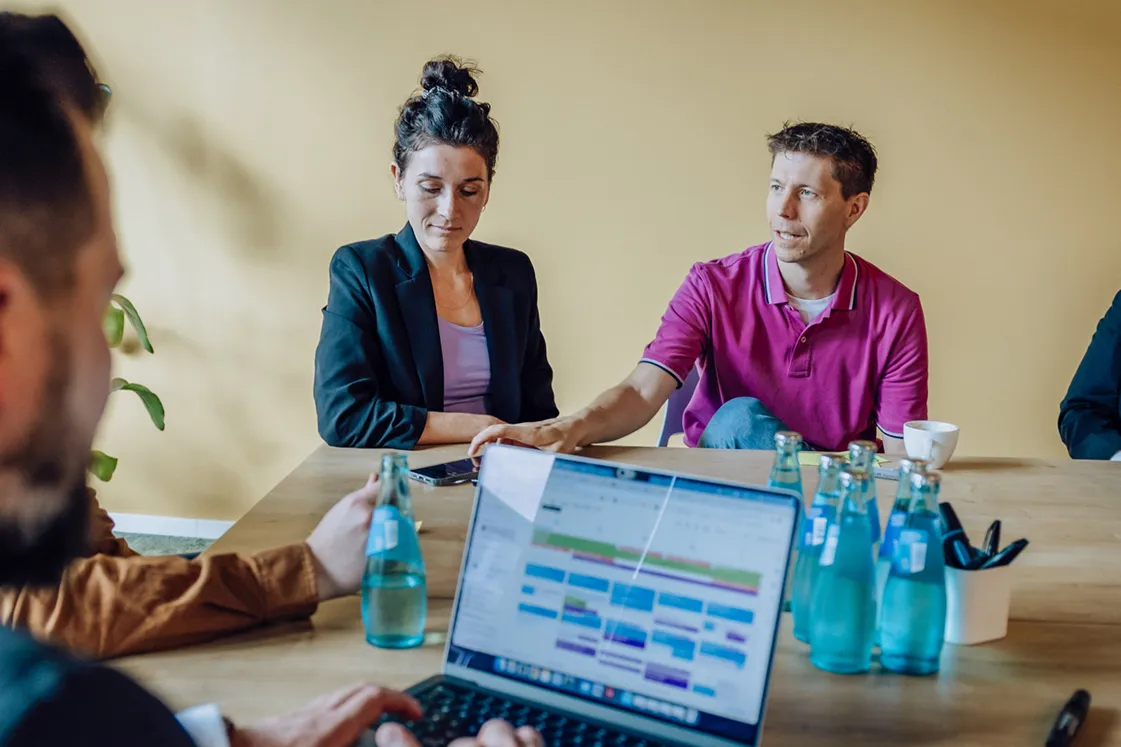 People having a meeting around a table with laptops and water bottles, against a yellow wall.