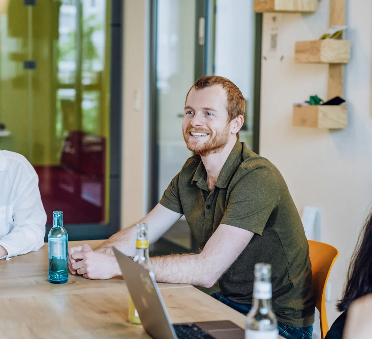 A group of people sitting around a table in an office setting, smiling and engaged in conversation. Bottles and a laptop are on the table.