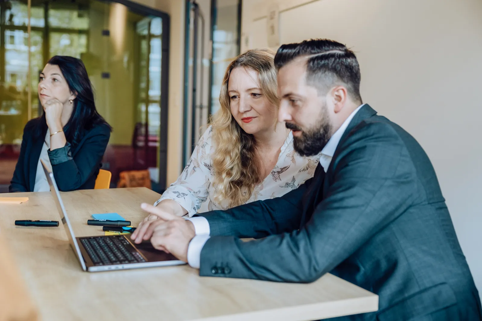 Three people in a meeting room, two focused on a laptop and one listening attentively.