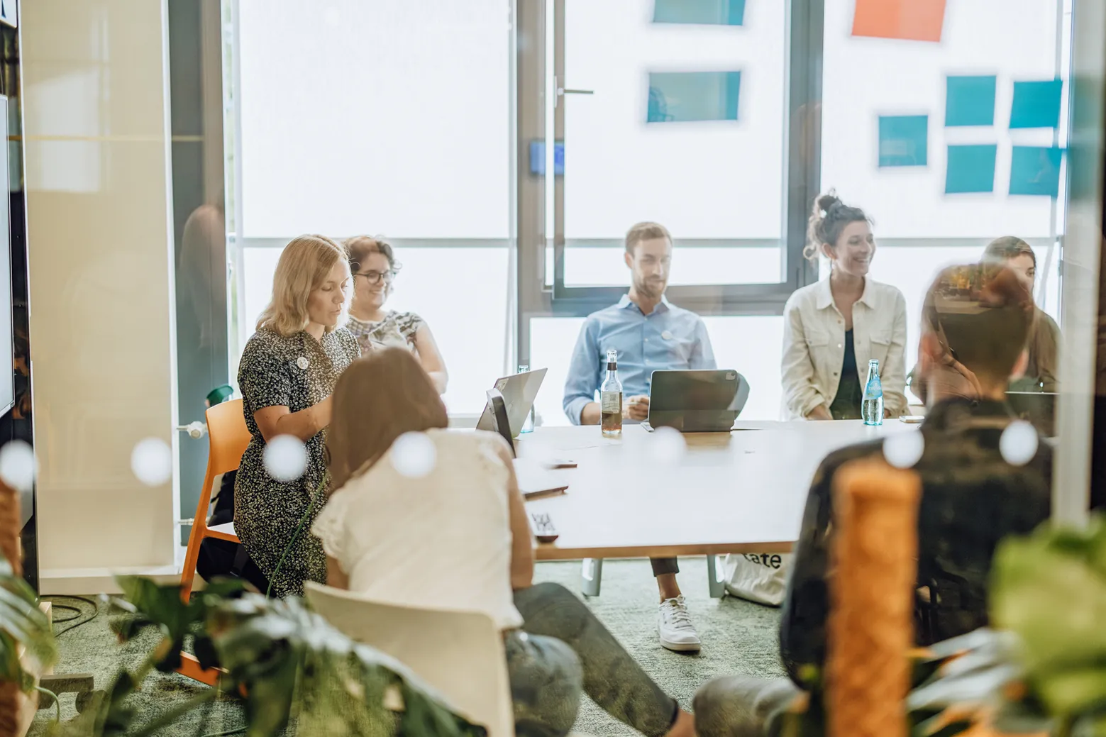 A group of people sitting around a table in a modern office setting, engaged in a meeting. Some have laptops open, and there are bottles of water on the table. The room has large windows and plants are visible in the foreground.