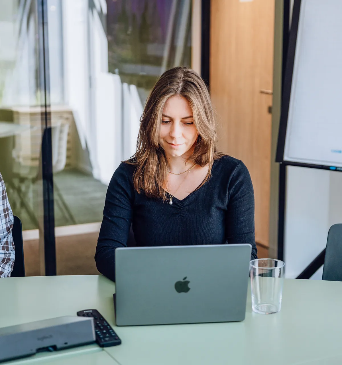 A woman working on a laptop at a green table in an office setting, with a man smiling beside her and a flipchart in the background.