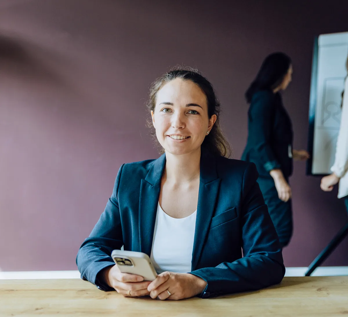 A woman in a blazer smiling while holding a smartphone, sitting at a table. In the background, two people are discussing something on a flipchart.