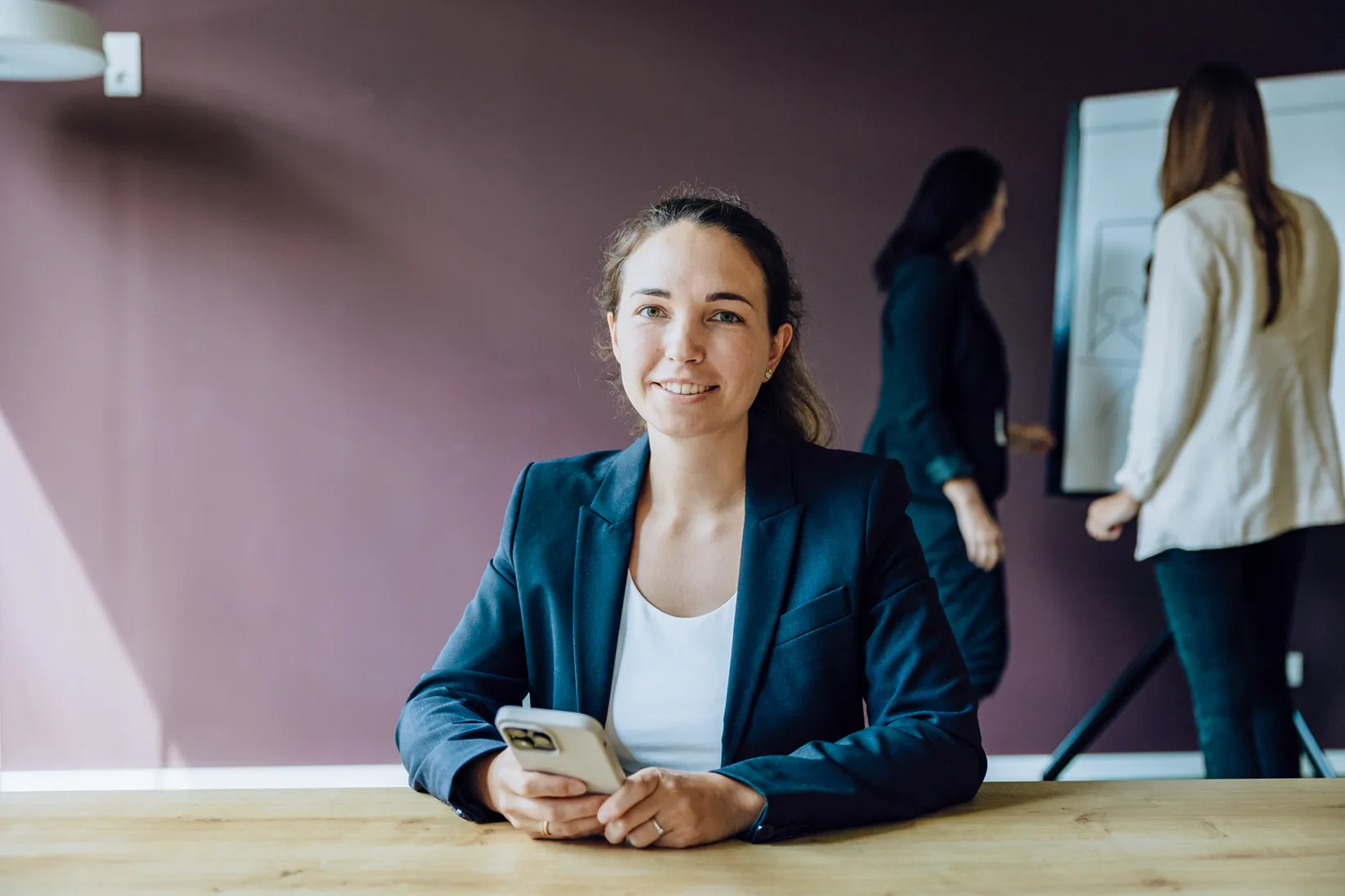 A woman in a blazer smiling while holding a smartphone, sitting at a table. In the background, two people are discussing something on a flipchart.